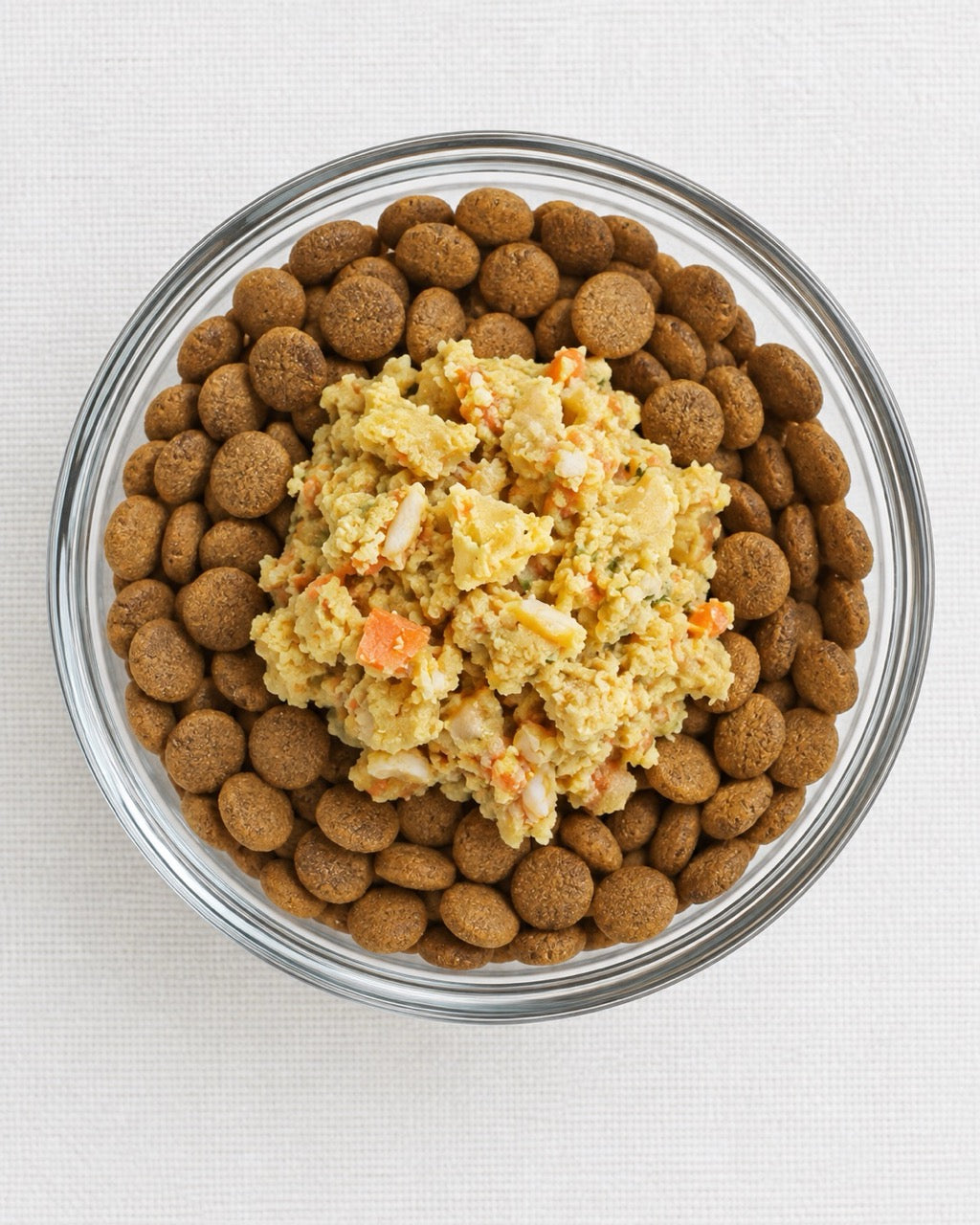 A clear glass bowl filled with dry brown kibble and a central scoop of  Furry Green fresh pet food with visible orange carrot pieces, photographed from above on a light fabric background.